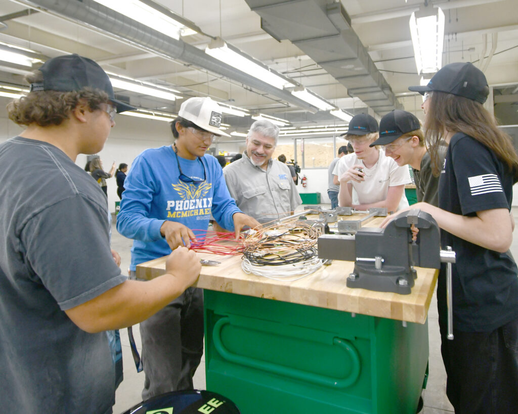 A man and several students smile as they watch another student demonstrate electrical work at a work table.