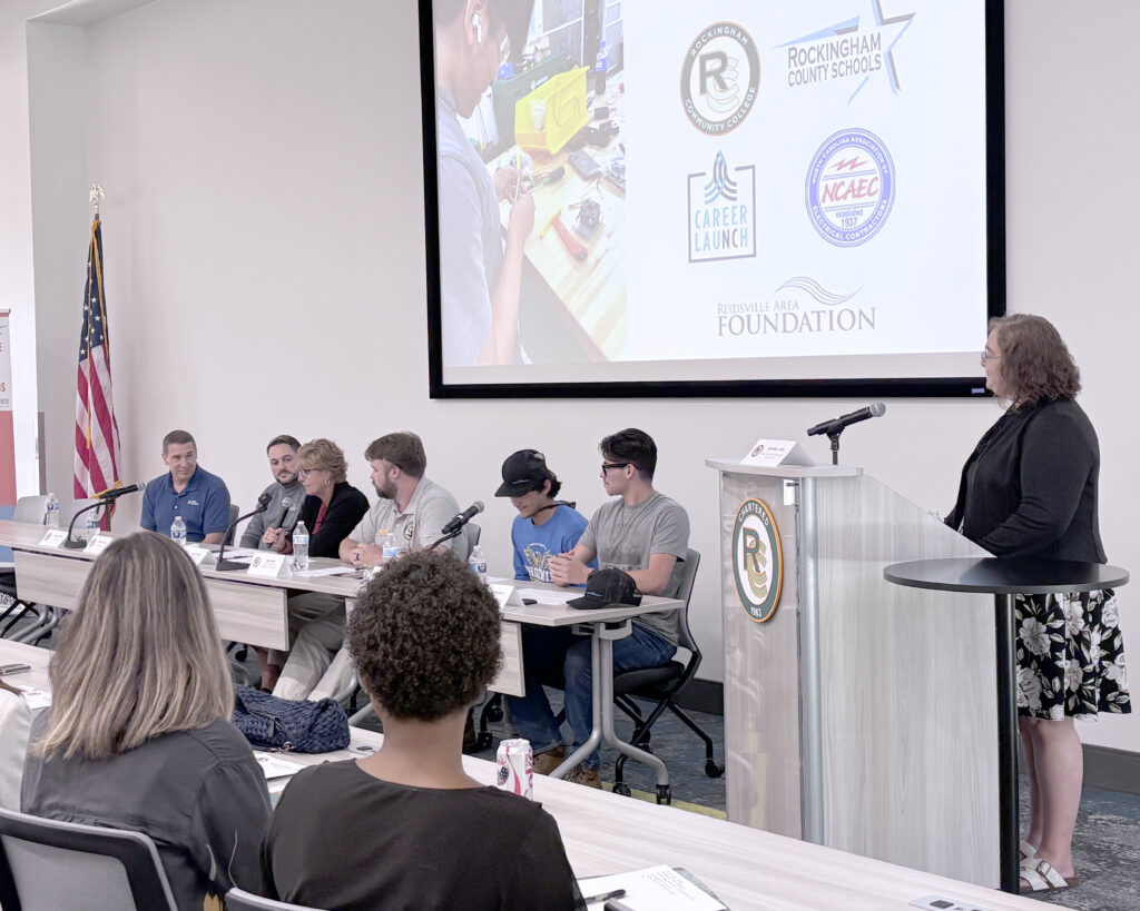 A woman stands at a podium, moderating a panel of six people.
