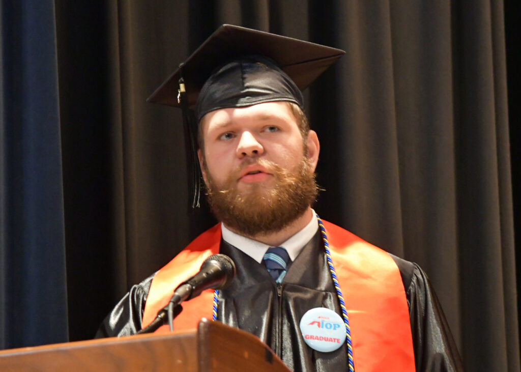 A man speaks at a podium, wearing a cap and gown and a RockATOP graduate button.