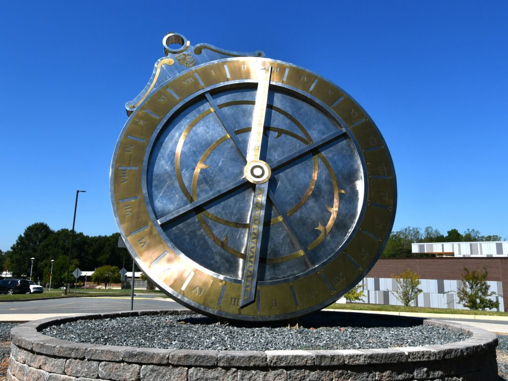 Large metal astrolabe sculpture with Roman numerals and brass accents mounted in a circular stone planter on a college campus under a clear blue sky.